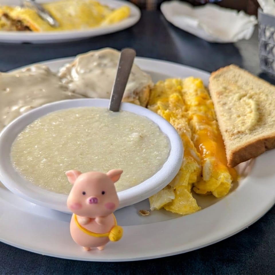 Breakfast plate with biscuits and gravy, scrambled eggs, toast, and a small pink pig figurine on a restaurant table.
