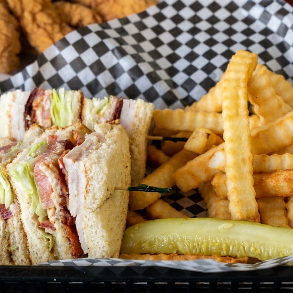 Basket of club sandwich bites with lettuce and bacon, crinkle-cut fries, pickle spear, and fried chicken tenders.